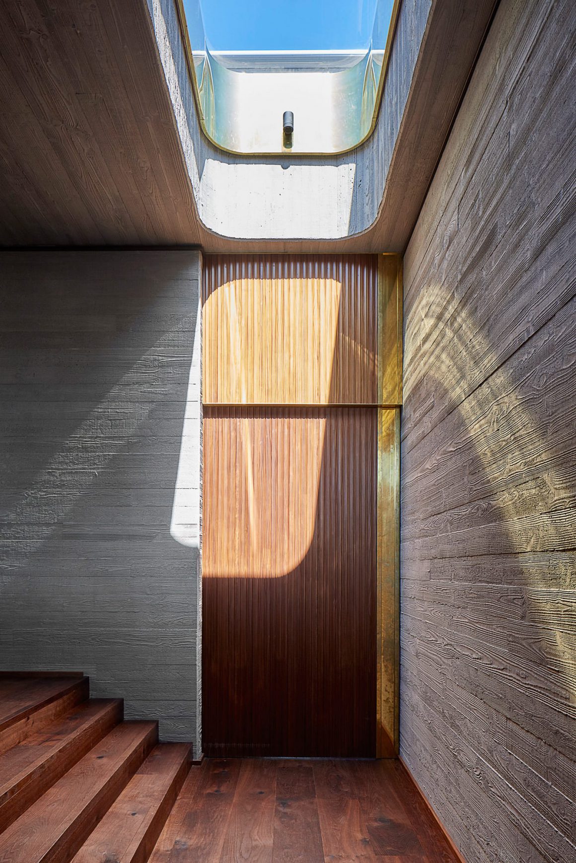 Porte d'entrée principale de la maison en bois bicolore brun et doré avec des lames fines et verticales qui contrastent avec les larges planches brut marron clair des murs. CP. Jack Lovel