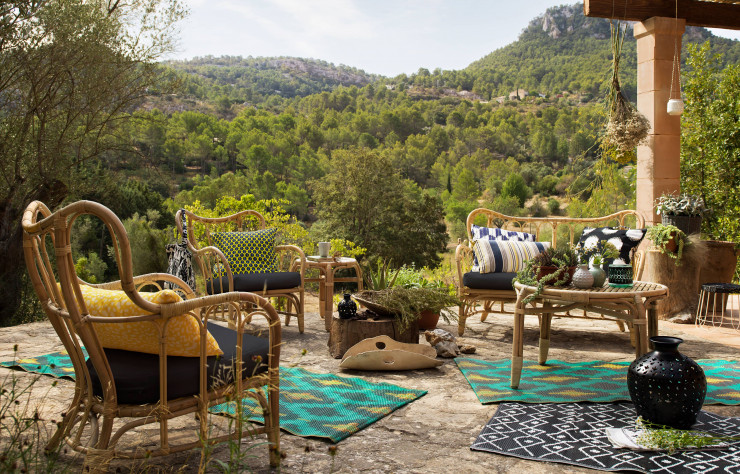 Salon de jardin en rotin avec des coussins et tapis dans les tons jaunes, bleus, blancs, noirs et verres pour que le salon s'intègre à la nature environnante. 